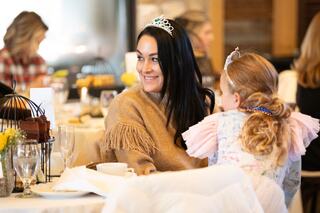 A woman in a cozy sweater and tiara smiles while a girl in a colorful dress leans in, surrounded by a festive dining setting.