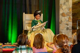 A princess in a green and yellow gown sits in a chair, reading to captivated children in a festive, colorful setting.