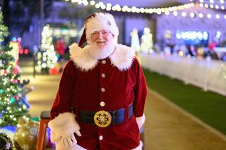 A cheerful man in a classic red Santa Claus outfit, surrounded by festive lights and decorations, exudes holiday spirit.