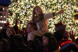 A joyful crowd celebrates around a beautifully lit Christmas tree, with a girl on a man’s shoulders, reaching out to the festive scene.