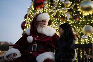 A joyful Santa in a red suit chats with a young girl, framed by a beautifully lit Christmas tree adorned with ornaments.
