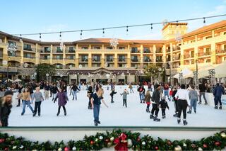 A festive outdoor ice skating rink is bustling with families and children, surrounded by holiday decorations and a charming building backdrop.