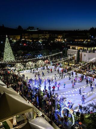 A festive outdoor ice skating rink is bustling with skaters under twinkling lights, surrounded by holiday decorations and glowing trees.