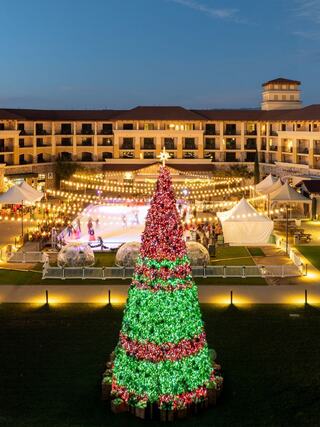 A vibrant Christmas tree adorned with lights stands near an outdoor ice rink, surrounded by festive decorations and a lively crowd.