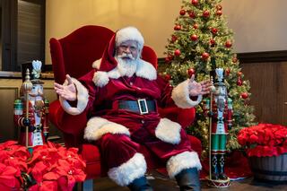 A cheerful Santa Claus in a red suit sits on a large chair, surrounded by festive decorations, including a Christmas tree and nutcrackers.