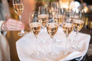 A tray holds several elegant champagne flutes filled with sparkling wine, with a hand reaching for one glass.