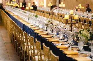 An elegant long dining table is set with golden chairs, glassware, and neatly arranged plates, illuminated by soft candlelight.