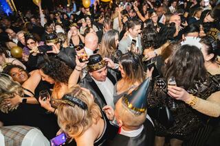 A lively crowd celebrates, dressed in stylish attire and party hats, with drinks in hand, amidst a festive atmosphere filled with laughter.