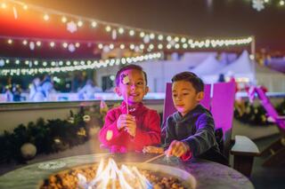 Two cheerful children roast marshmallows by a glowing fire pit, surrounded by festive lights and a cozy winter atmosphere.