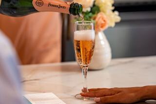A hand reaches out as champagne is poured into a flute, surrounded by soft flowers and a cozy setting, conveying a celebratory atmosphere.