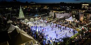 A bustling outdoor ice skating rink surrounded by festive lights, a towering Christmas tree, and a vibrant crowd enjoying the holiday spirit.