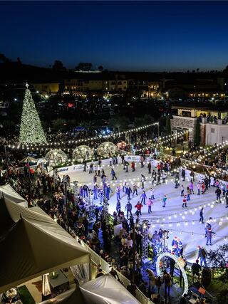 A festive ice skating event with twinkling lights, a large Christmas tree, and crowds enjoying the holiday spirit at twilight.