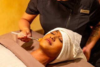 A woman receives a facial treatment while relaxing on a massage table, with a therapist applying a mask using a brush.