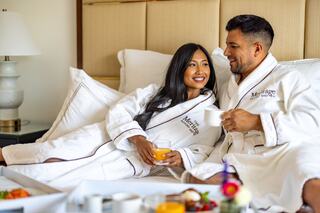 A couple in cozy robes enjoys a relaxing morning together in bed, sharing smiles and breakfast with a warm atmosphere.