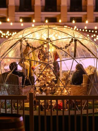 A cozy, illuminated igloo is filled with festive decorations and people enjoying a celebratory atmosphere, flanked by strings of lights.
