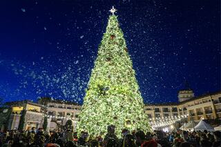 A beautifully decorated Christmas tree shines brightly, surrounded by joyful crowds and falling artificial snow against a twilight sky.