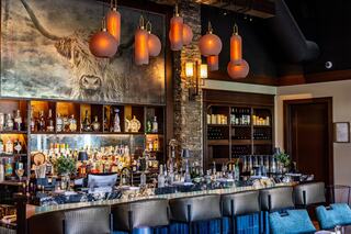 A stylish bar features a vibrant display of spirits, chic hanging lights, and a striking wall mural of a Highland cow, creating a modern atmosphere.