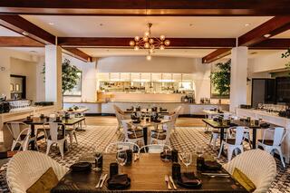 A stylish restaurant features wooden beams, modern decor, patterned flooring, and elegant tables set for dining. Greenery adds a fresh touch.