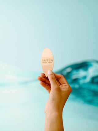 A hand holds a wooden tag inscribed with "PASEA HOTEL & SPA" against a serene blue backdrop with gentle wave patterns.