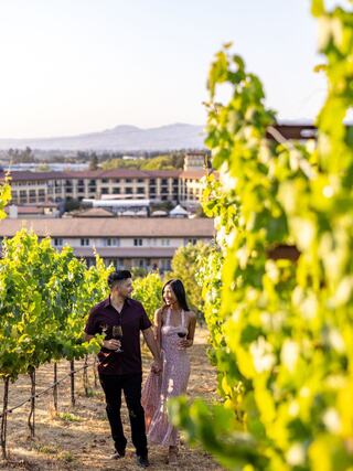 A couple strolls hand in hand through a vineyard, enjoying wine and the scenic view of rolling hills and a charming building in the background.