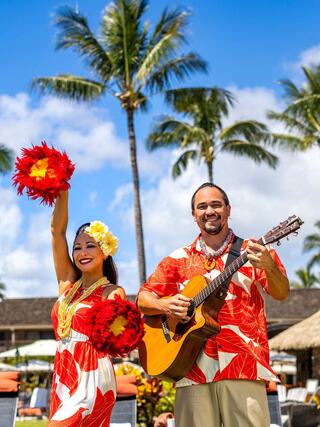A joyful duo performs outdoors, dressed in vibrant Hawaiian attire, with lush palm trees and a sunny blue sky in the background.