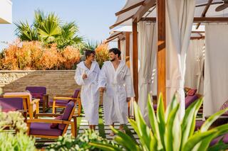 A couple in white robes strolls hand-in-hand through a relaxing outdoor spa area, surrounded by lush greenery and inviting lounge chairs.