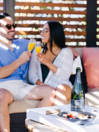 A couple enjoys mimosas on a cozy patio, sharing smiles and laughter, with a bottle of champagne and a fruit platter on the table.