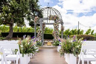 A charming outdoor wedding altar adorned with flowers, surrounded by white chairs and lush greenery, set against a picturesque vineyard backdrop.
