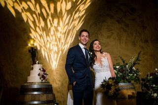 A smiling couple stands together in a warm venue, surrounded by floral arrangements and a wedding cake, illuminated by soft lighting.
