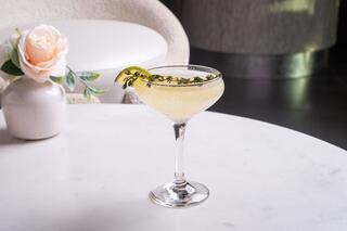 A delicate cocktail garnished with a lemon twist and herbs sits elegantly on a marble table beside a soft pink rose in a vase.