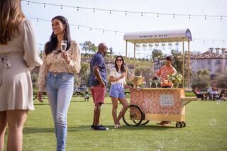 A vibrant outdoor scene with friends enjoying drinks at a charming cart serving bubbles and bites, surrounded by greenery and string lights.