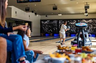 A child prepares to bowl while friends enjoy snacks and watch, with vibrant decor and screens enhancing the lively atmosphere.