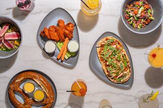 A variety of tasty dishes including wings, flatbread, pretzels, and fresh salads, accompanied by vibrant drinks, arranged elegantly on a table.