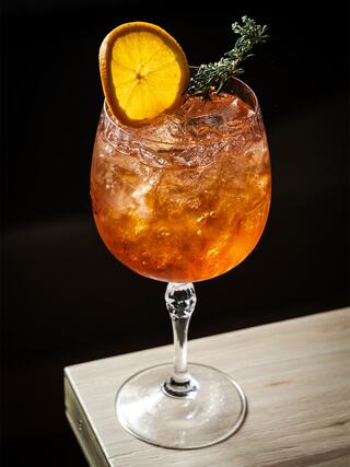 A refreshing cocktail with ice, garnished with a slice of orange and a sprig of thyme, served in an elegant glass.