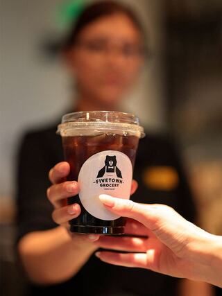 A barista hands over a cup of iced coffee branded with a logo from a grocery store, while another hand reaches to receive it.