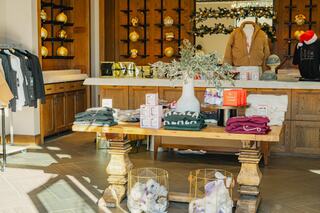 A cozy retail space features a wooden table with clothing items and decorative gifts. Stylish apparel hangs nearby, creating a warm atmosphere.