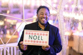 A man smiles brightly while holding a festive sign that reads "NOEL," set against a backdrop of warm lights and holiday decor.