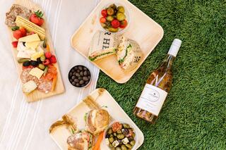 A picnic spread features sandwiches, cheese, fresh fruits, olives, chocolate, and a bottle of wine, all set on green grass.