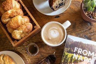 Croissants in a wooden basket, a creamy latte, a slice of cake, and a magazine on a rustic table create a cozy food scene.