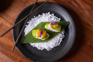 Two sushi pieces topped with golden uni, garnished with green leaves and a purple flower, served on a bed of coarse sea salt.