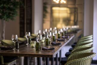 A long wooden dining table is elegantly set with glasses, napkins, and vases, accompanied by stylish green chairs in a warm atmosphere.