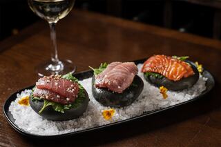 A stylish platter features three types of sushi atop black stones, garnished with greens and flowers, alongside a glass of white wine.