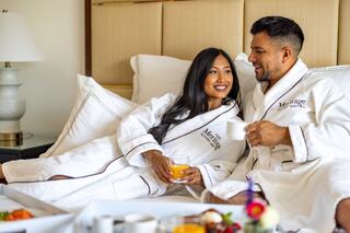 A couple in cozy robes relaxes together in bed, enjoying drinks and breakfast, sharing smiles in a serene, intimate setting.