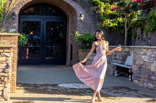 A woman in a floral dress twirls joyfully, holding a drink, in a charming outdoor space with brick walls and greenery.