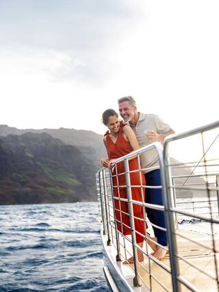 A joyful couple enjoys a scenic moment on a boat, embracing by the railing with mountains and ocean in the backdrop.