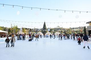 Outdoor ice rink with people skating, surrounded by festive decor and a large Christmas tree. String lights and snowflakes hang above, creating a joyful atmosphere.