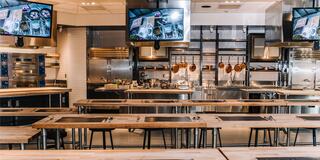 A modern, spacious kitchen with wooden tables, overhead screens, and well-organized cooking stations, exuding a professional atmosphere.