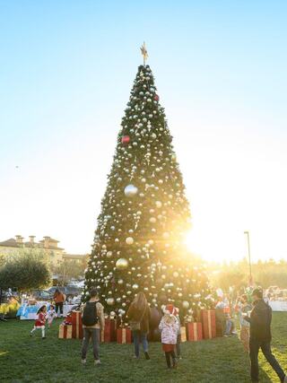 A large, decorated Christmas tree stands tall against a sunset backdrop, with people in festive attire gathered around and presents at its base.