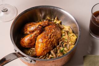 Roasted chicken glistens atop a bed of creamy noodles and sautéed mushrooms, all served in an elegant copper pot. A glass of dark beverage nearby.