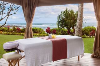 A serene outdoor spa setup features a massage table with towels, overlooking a tranquil ocean view and vibrant tropical greenery.